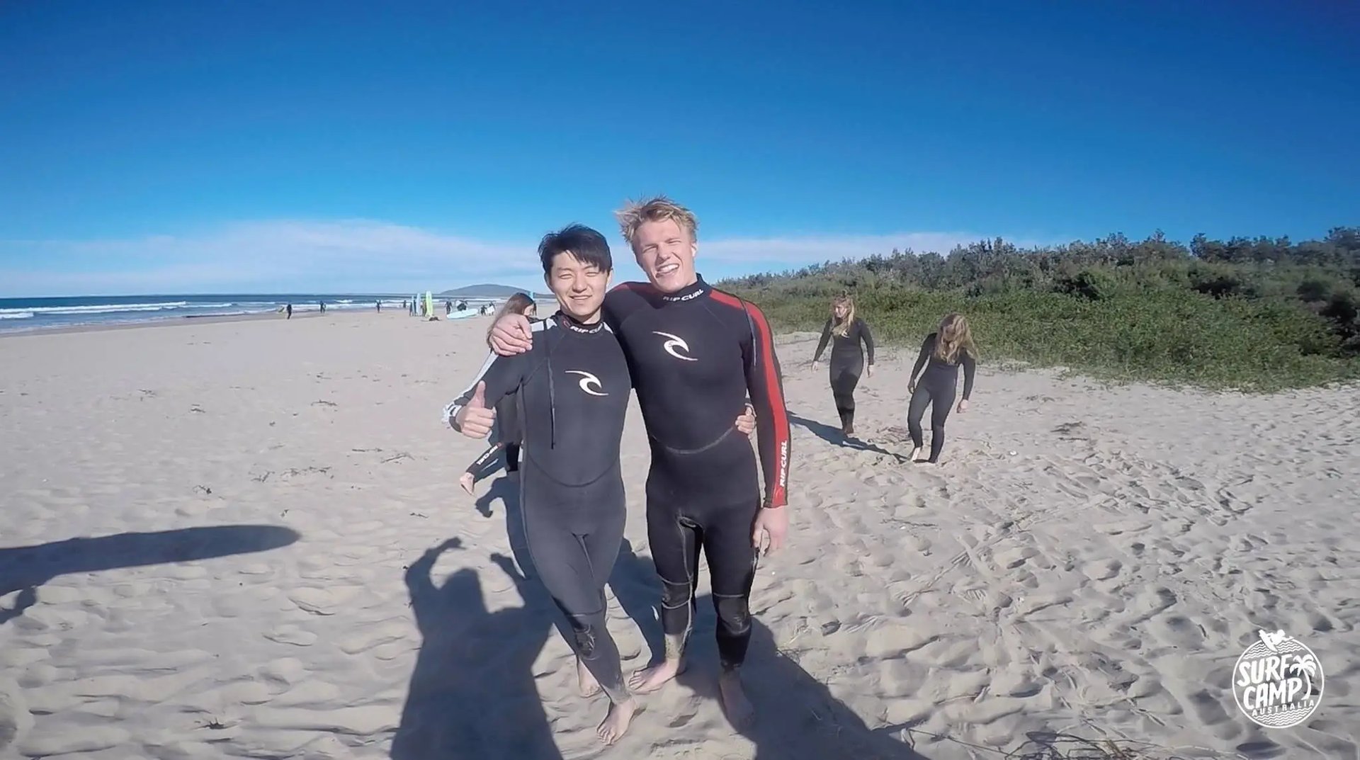 Zwei Studenten in Neoprenanzügen stehen lächelnd am Strand, umgeben von Sand und Meer.