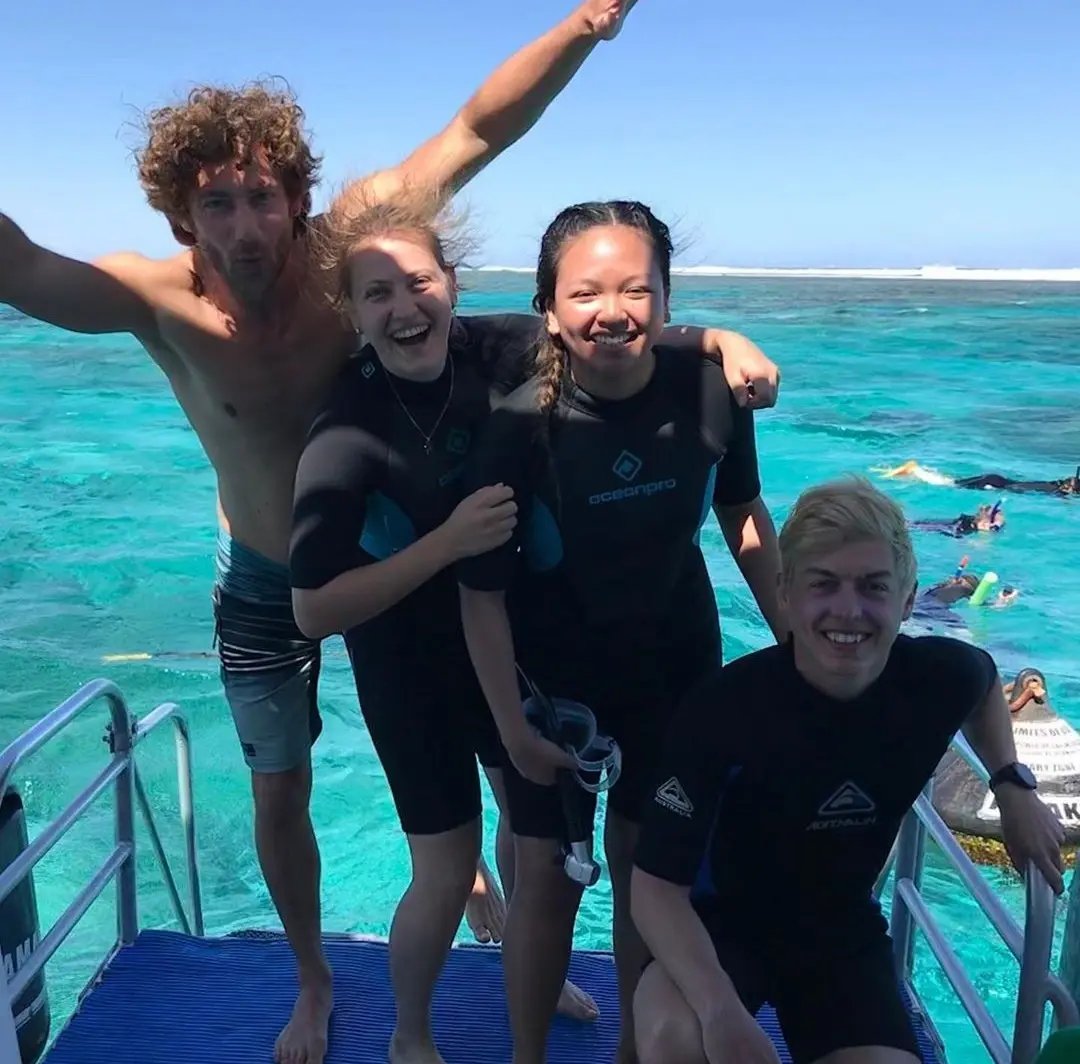 ruppe von vier Studierenden in Taucheranzügen lacht auf einem Boot am Meer. 