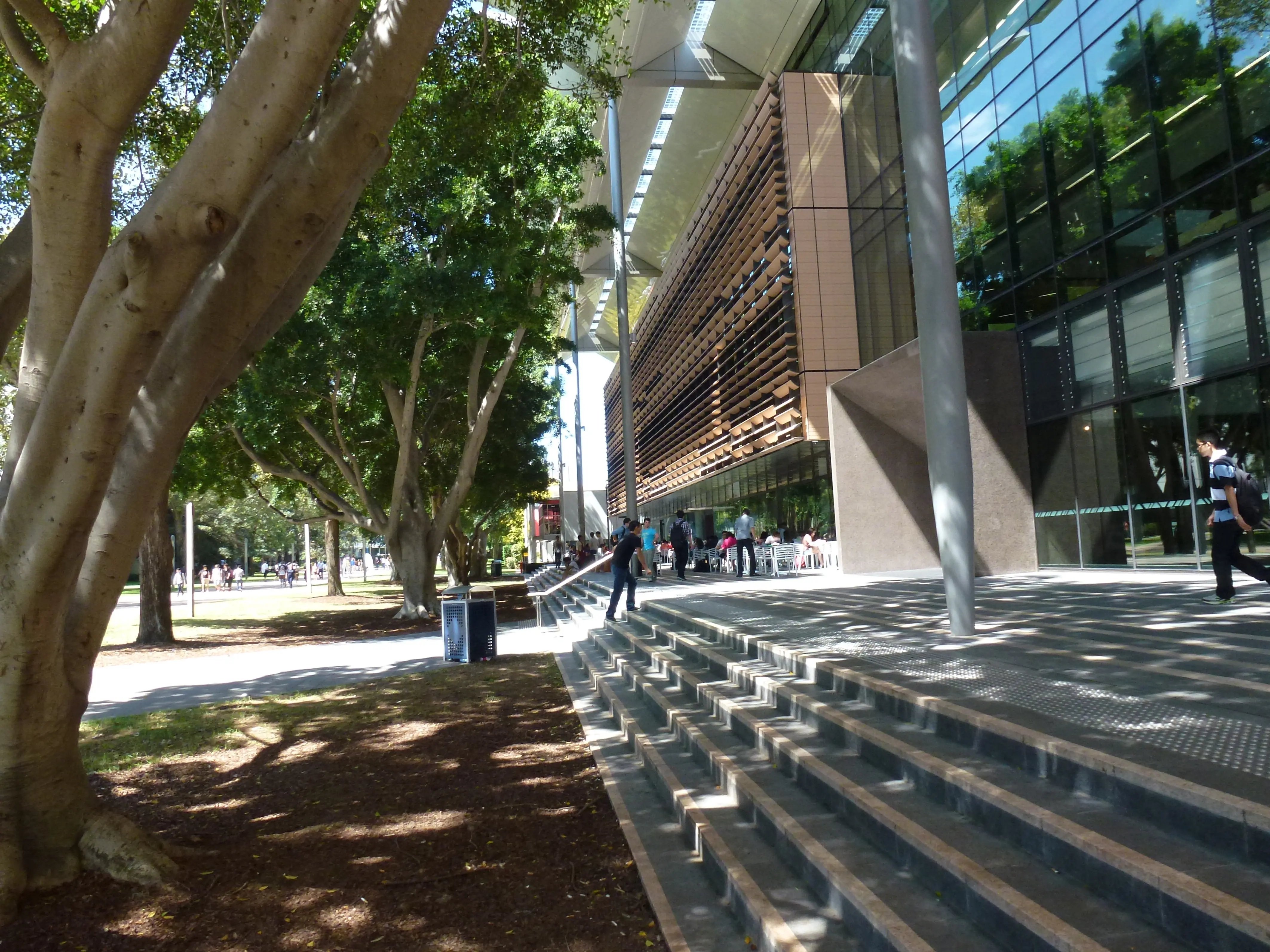 Eingangsbereich eines modernen Gebäudes auf dem Campus der University of New South Wales mit breiten Treppenstufen, schattenspendenden Bäumen und Studierenden im Hintergrund.