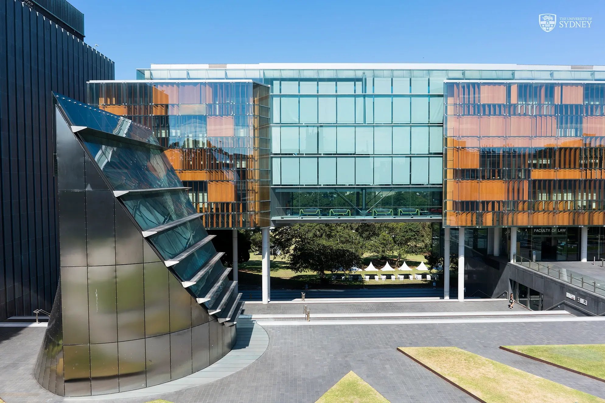 Moderne Glasfassade eines Universitätsgebäudes mit grünen Flächen und architektonischen Elementen im Vordergrund.