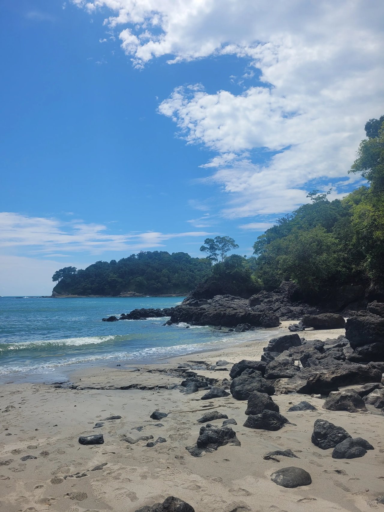 Ein naturbelassener Sandstrand mit dunklen Felsen, sanften Wellen und grüner Vegetation im Hintergrund.