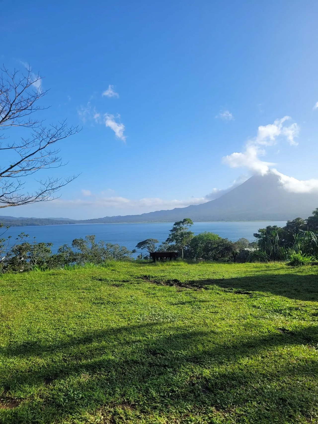 Eine grüne Wiese mit Blick auf den ruhigen Arenalsee und den in Wolken gehüllten Vulkan Arenal.