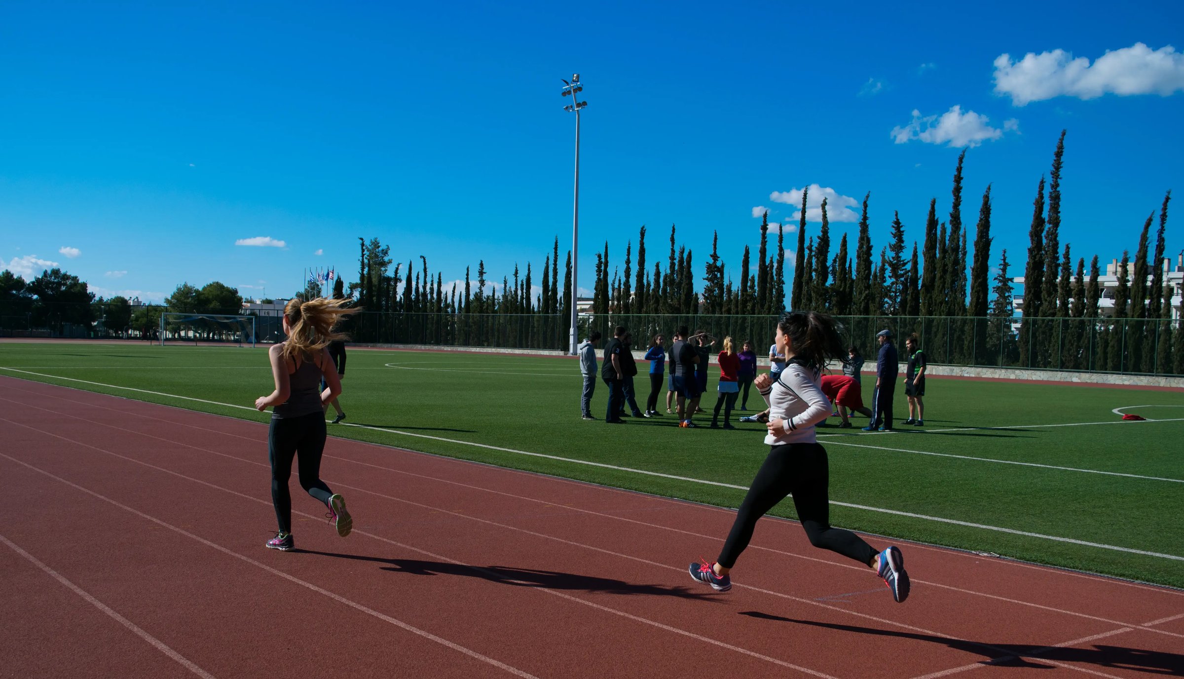 Zwei Studentinnen laufen auf einer Sportbahn unter blauem Himmel. 