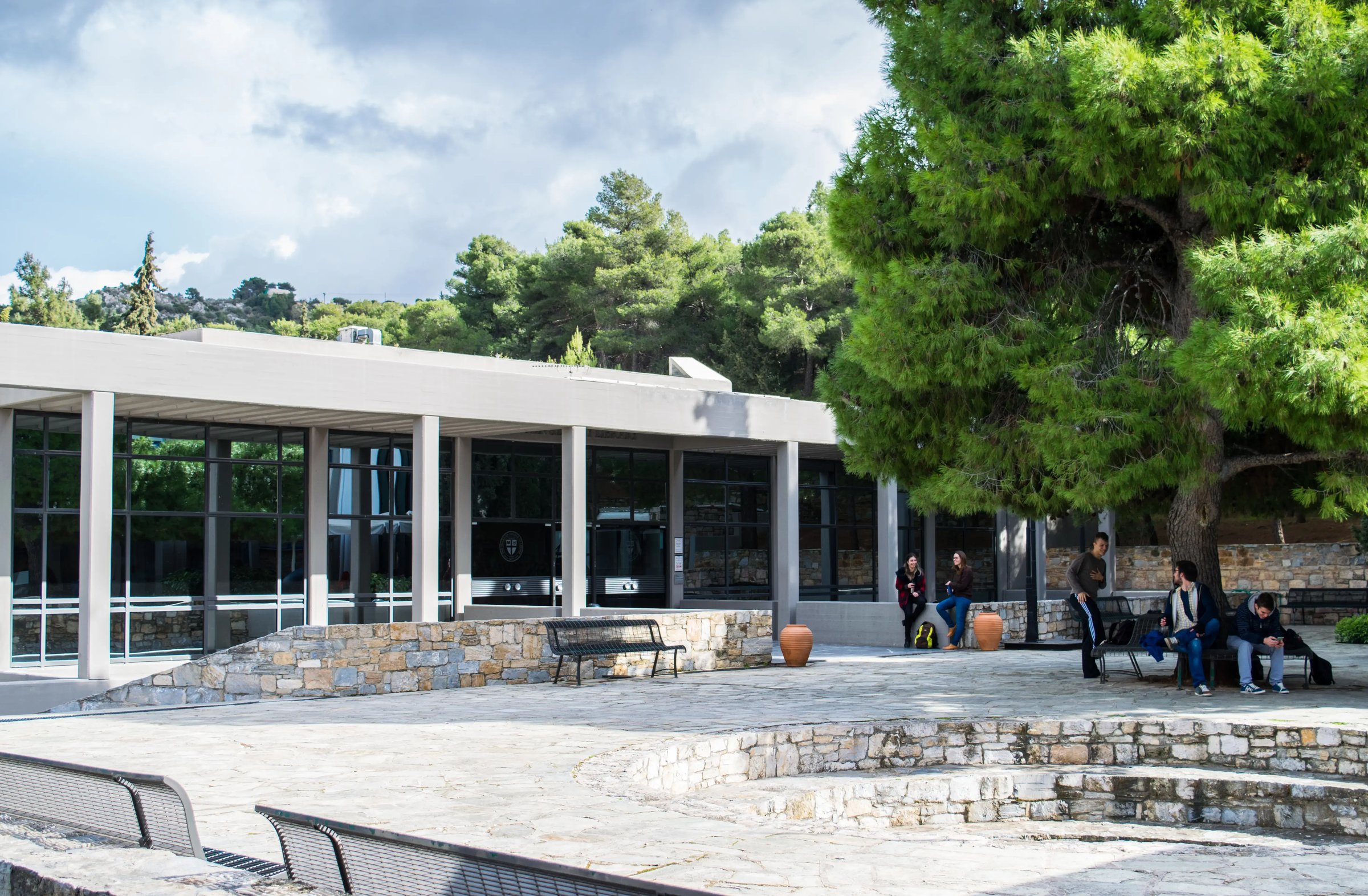 Moderner Campus American College of Greece mit Studierenden auf dem sonnigen Außengelände, umgeben von Pinienbäumen.