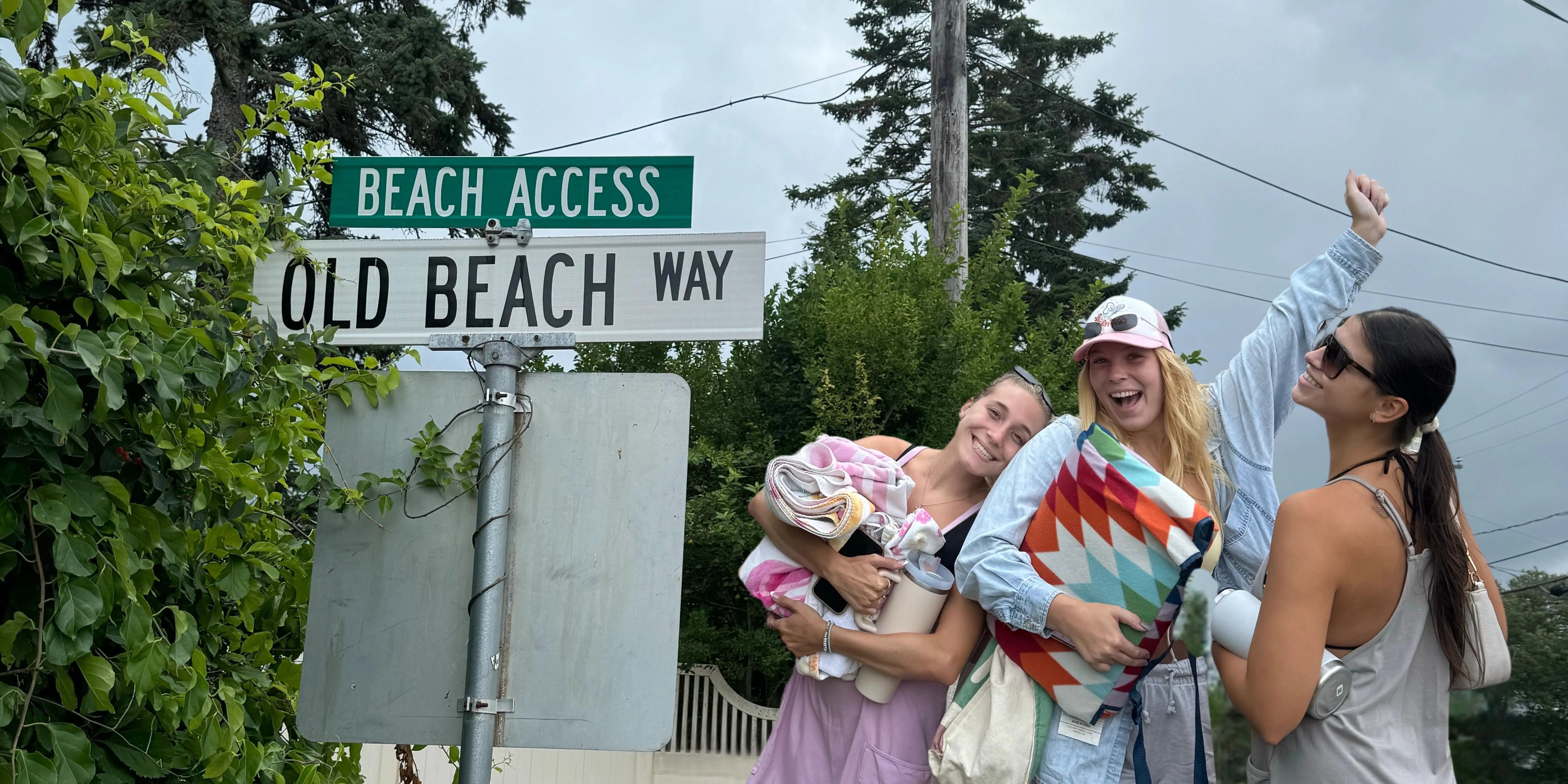 Drei Studentinnen lächelnd und in Sommerkleidung vor einem "Beach Access" Straßenschild.