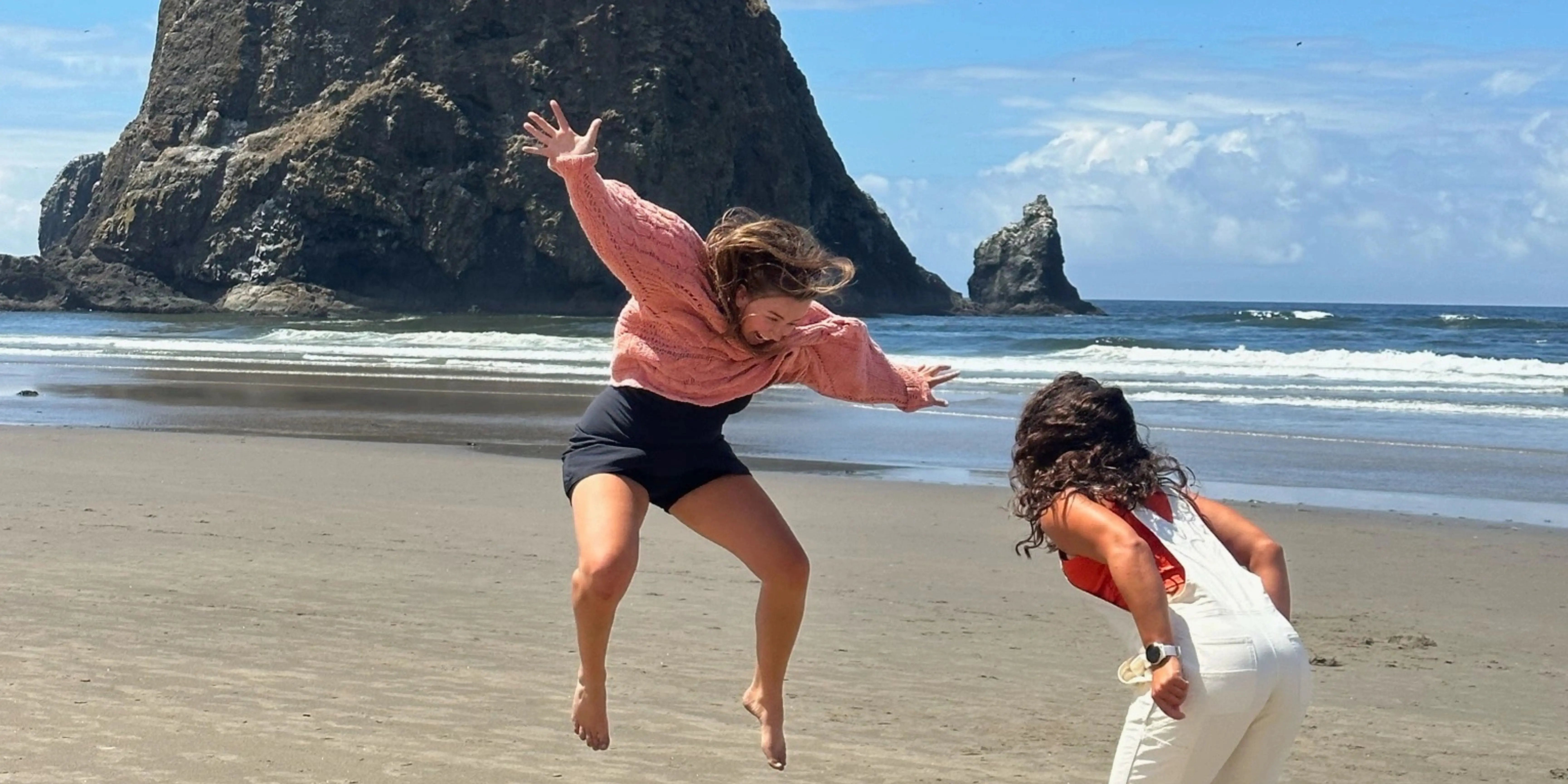 Zwei Frauen am Strand vor einem großen Felsen, eine springt freudig in die Luft, blauer Himmel im Hintergrund.. 