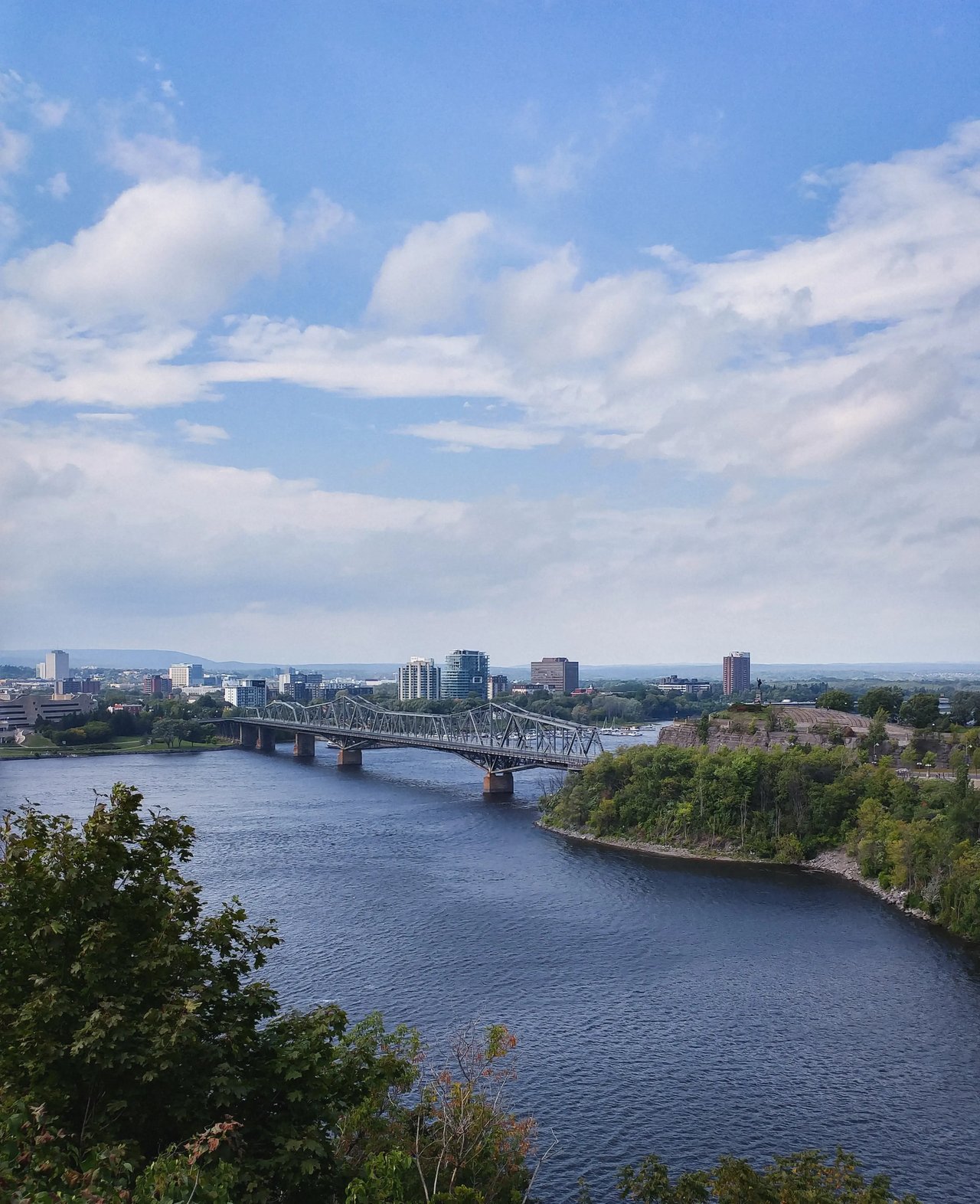 Blick auf den Ottawa River und die Skyline von Ottawa mit Brücken und grünen Bäumen.