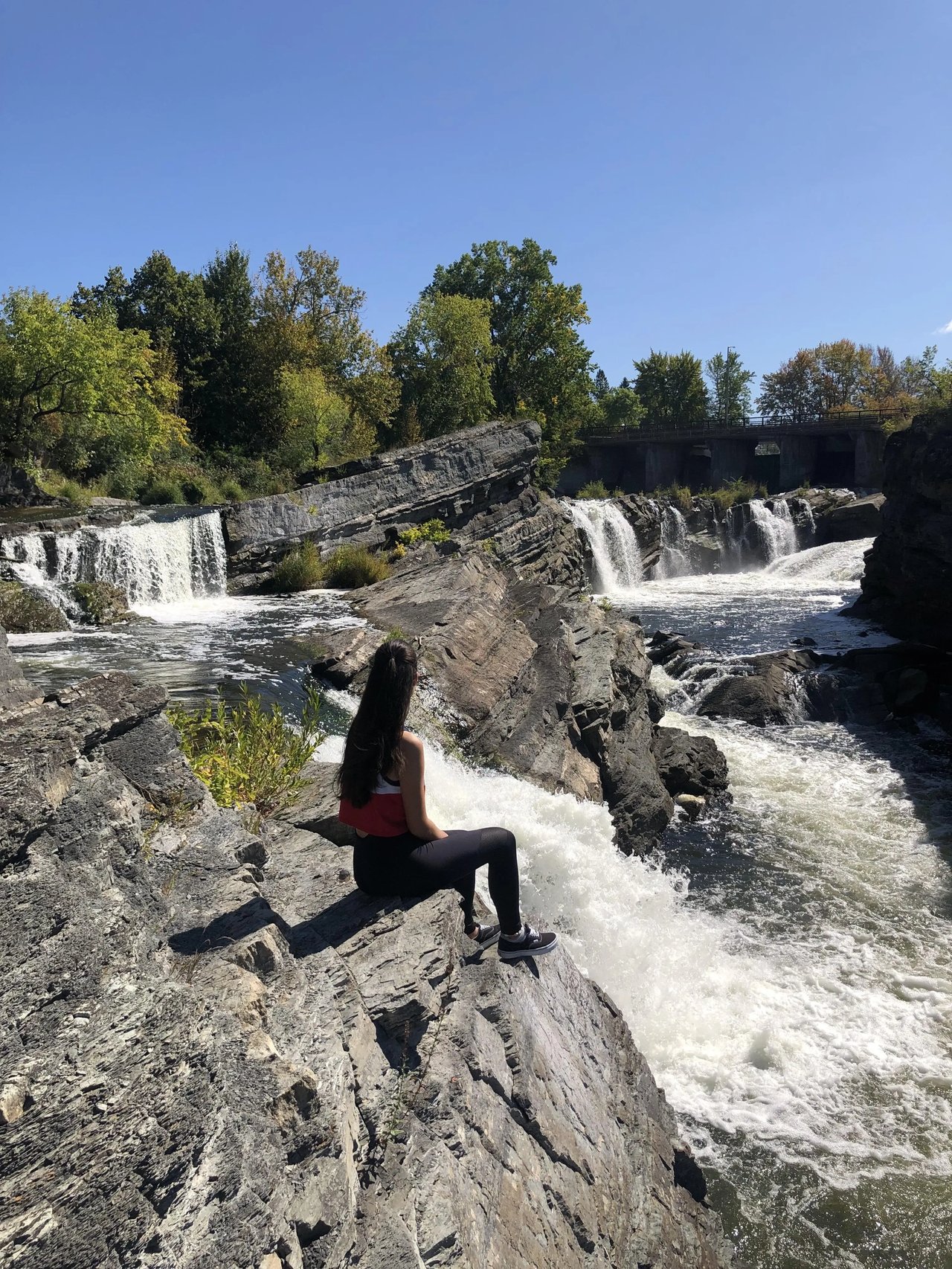 Studentin sitzt an einem Flussufer mit Wasserfall bei sonnigem Wetter in Ottawa, Kanada.