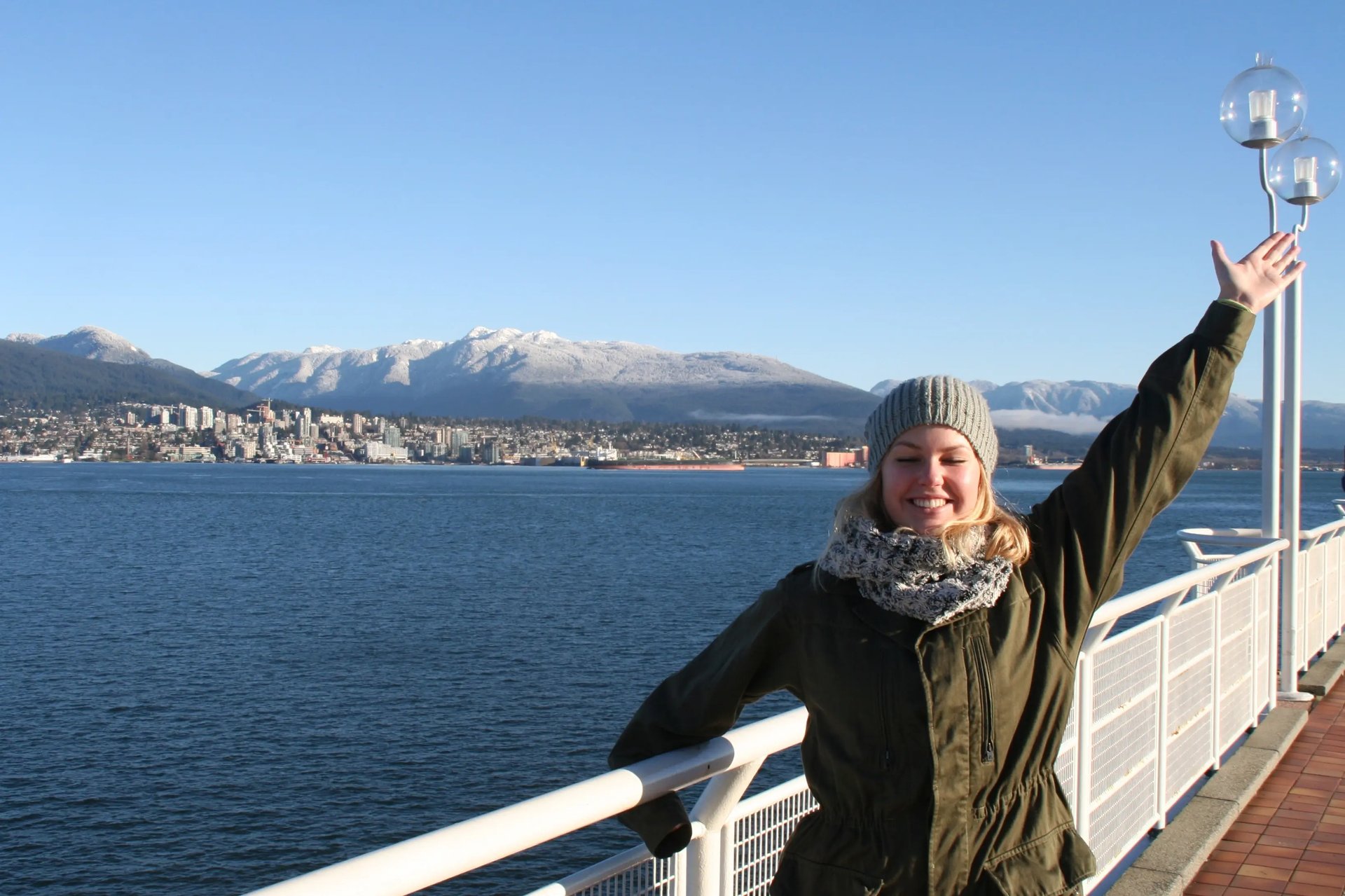 Studentin der University of Victoria mit strahlendem Lächeln auf einer Fähre, im Hintergrund die Skyline von Vancouver und schneebedeckte Berge.