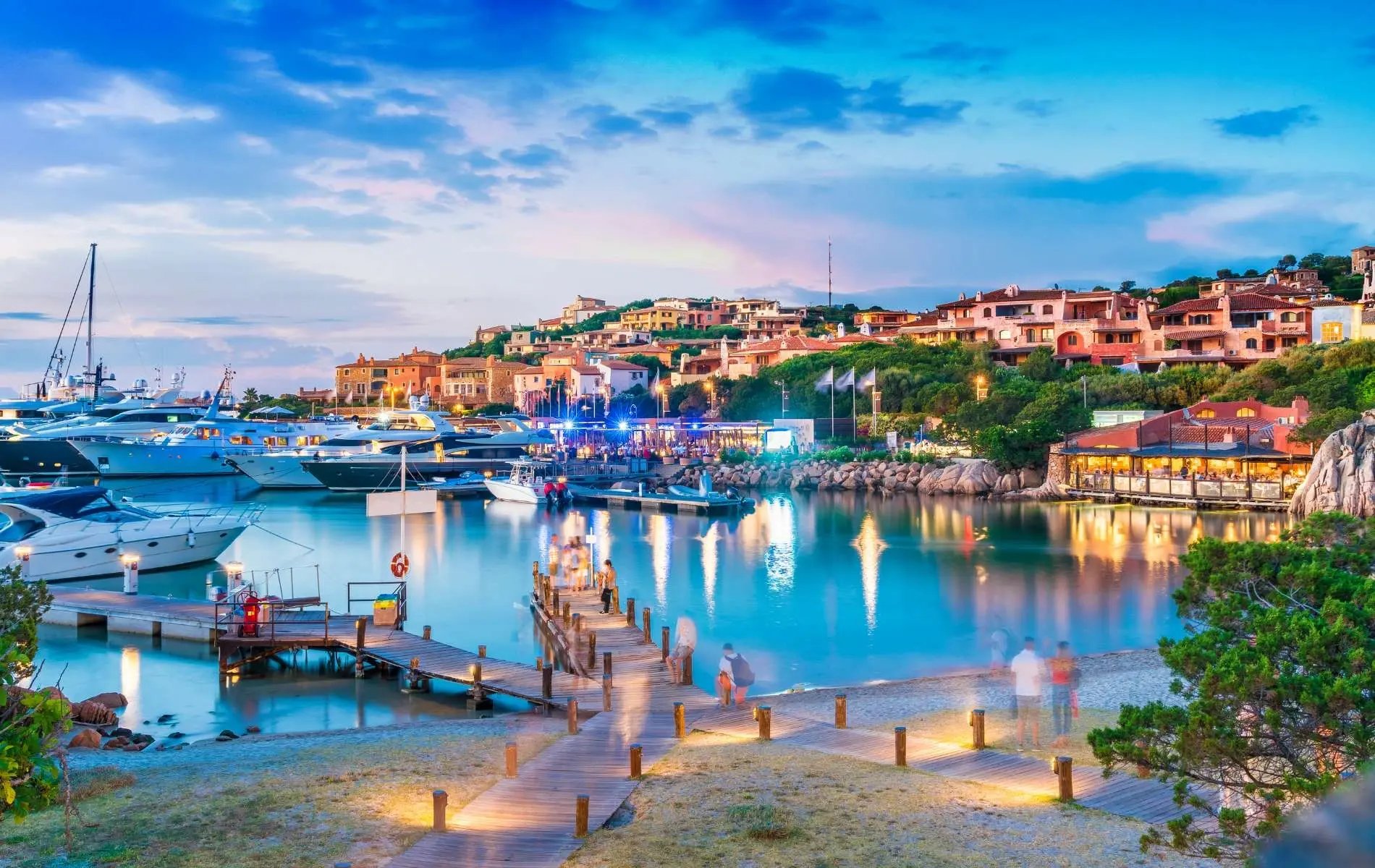 Hafen von Porto Cervo auf Sardinien, Italien, bei Dämmerung. Luxusyachten liegen im ruhigen, türkisblauen Wasser, während bunte Lichter die Hafenpromenade und die mediterranen Gebäude erhellen.