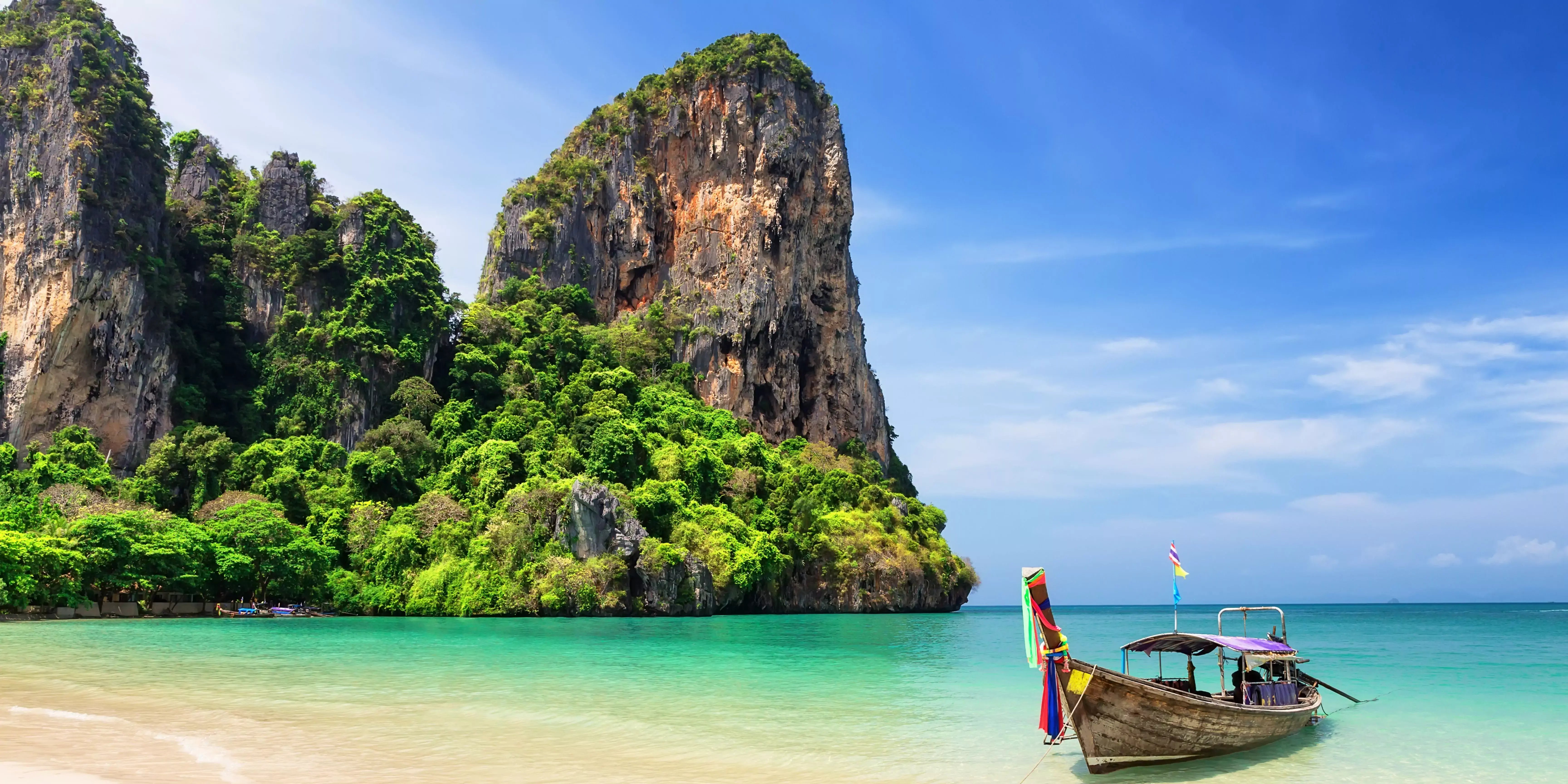 Tropische Küstenlandschaft in Thailand mit grün bewachsenen Felsen, blauem Himmel und klarem Wasser. Im Vordergrund ein traditionelles Longtail-Boot. 