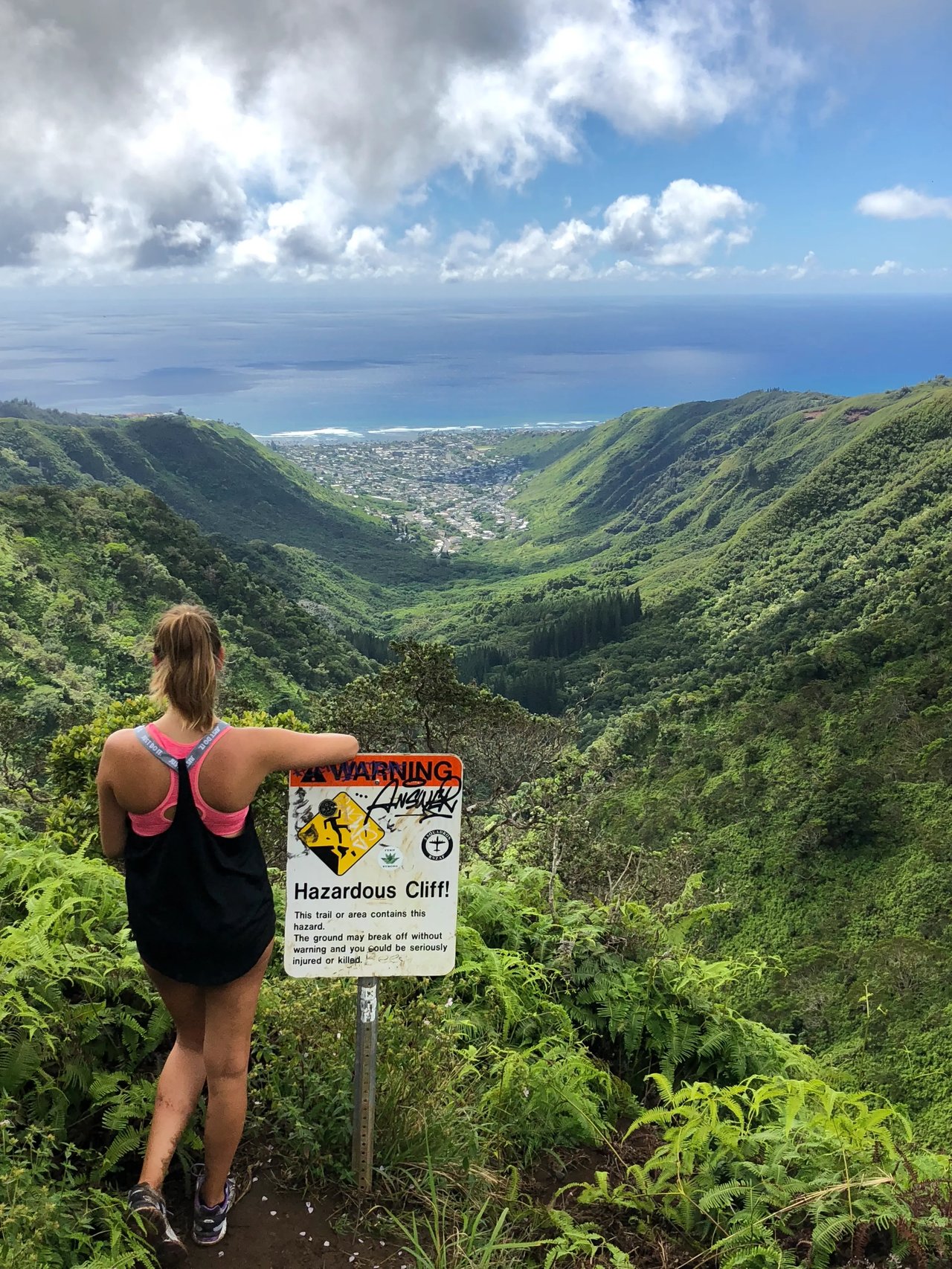 Aussicht auf grüne Berglandschaft und Küste von Oʻahu, Studentin der Hawaii Pacific University vor einem Warnschild auf einem Wanderweg.