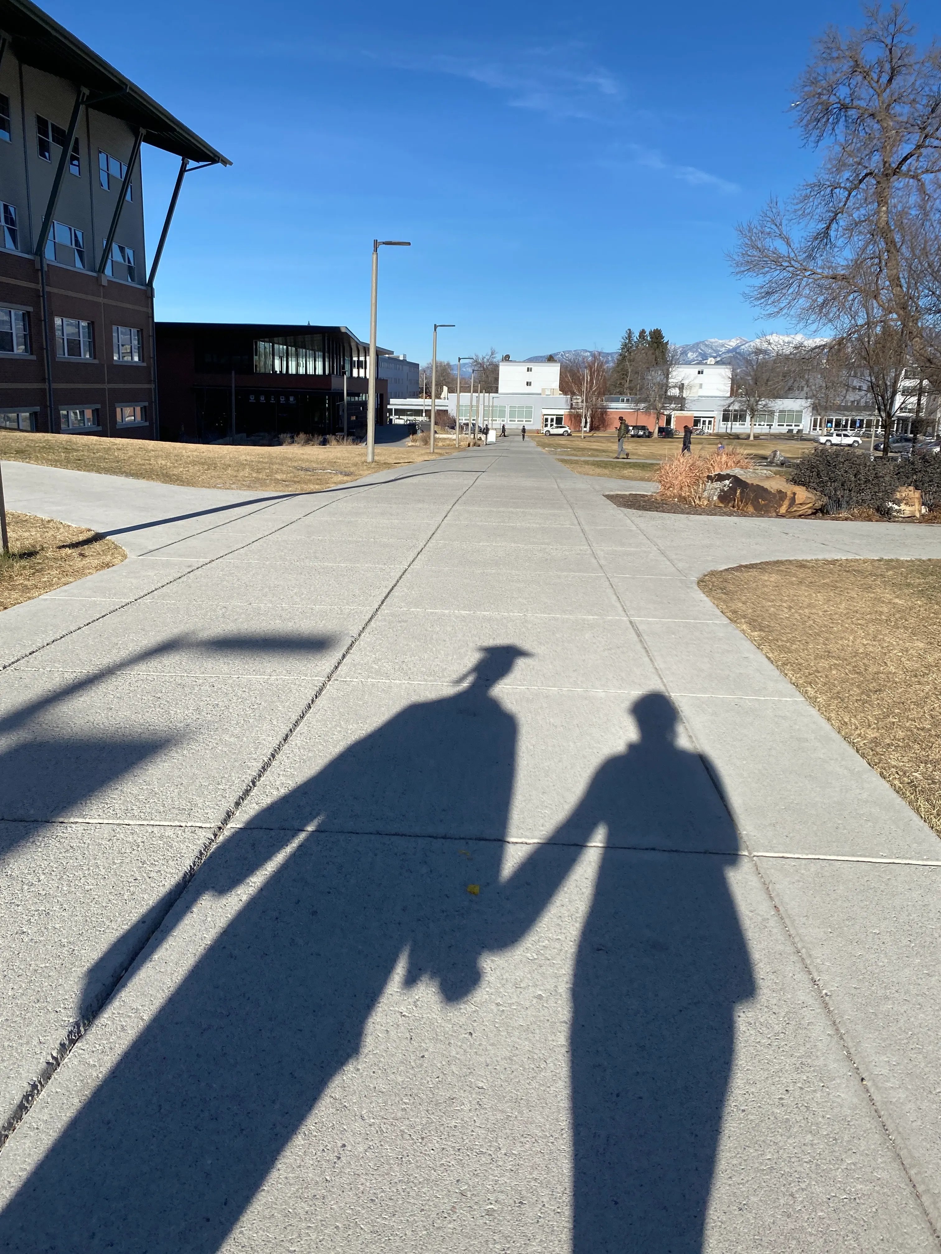 Schatten eines Absolventen mit Abschlusskappe und einer Begleitperson auf einem Campusweg.